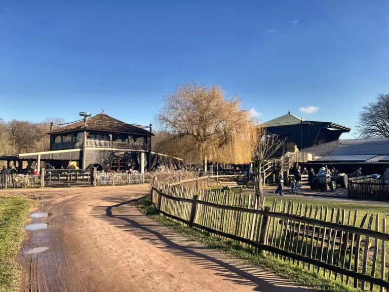 A charming view of the Amsterdamse bos featuring rustic buildings and a wooden fence along a dirt path under a bright blue sk