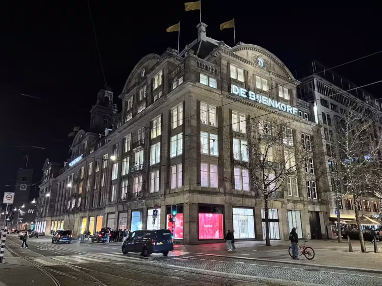 The illuminated façade of the Bijenkorf Amsterdam showcases its grandeur at night while shoppers mingle outside enjoying the