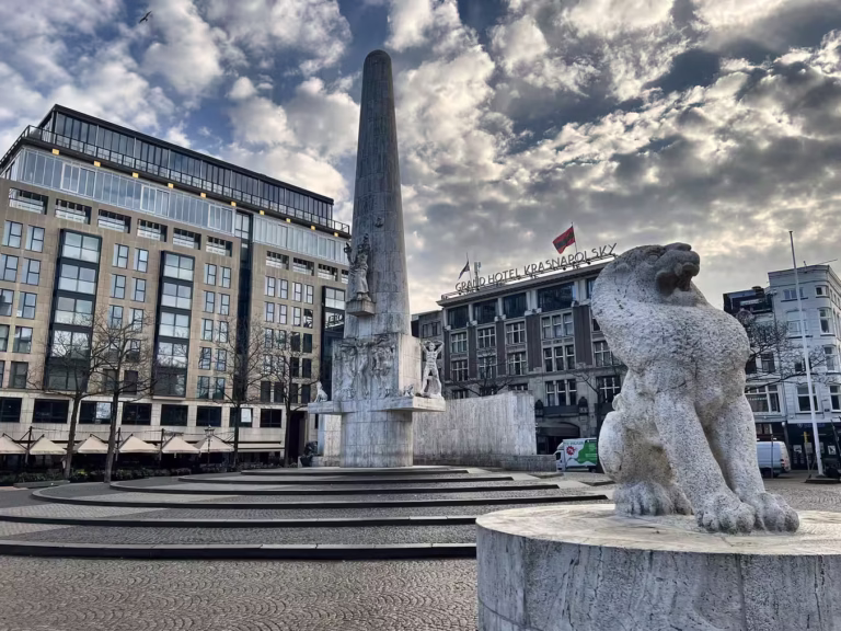 A lion statue stands in the foreground as the Dam square monument rises in the background against a cloudy sky