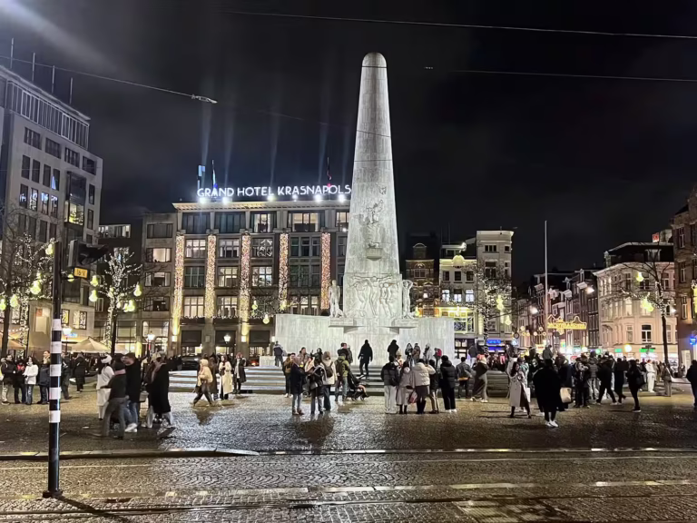 The lively Dam square features a prominent monument surrounded by crowds at night and illuminated buildings creating a vibran