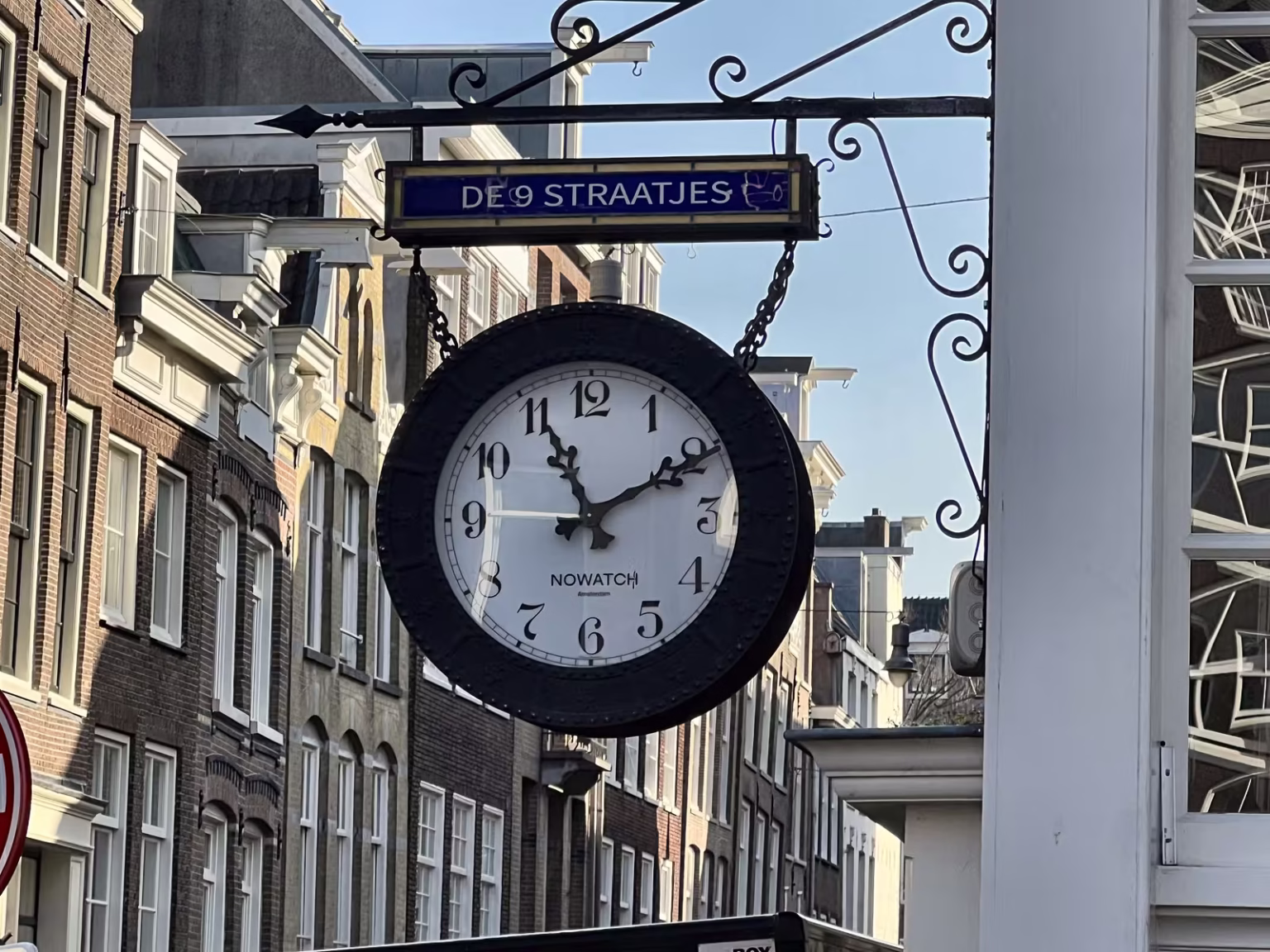 A stylish clock hangs from a sign pointing towards De Negen Straatjes with historical buildings lining the street