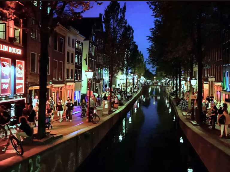 At night the colorful lights of bars and shops illuminate De wallen while people stroll along the canal's edge