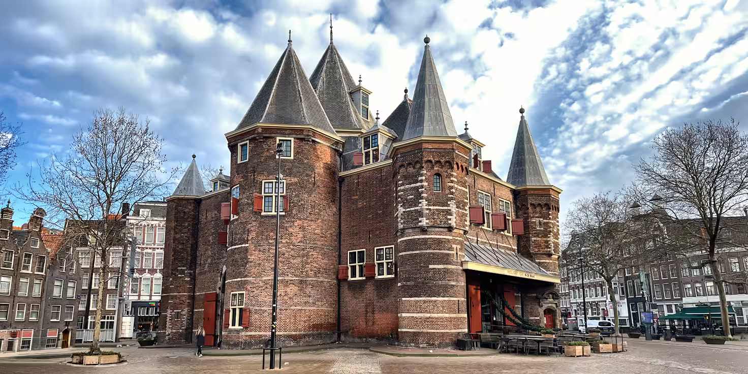 A historic brick building de Waag - with pointed towers stands in a square under a blue sky inviting visitors to discover its rich history,