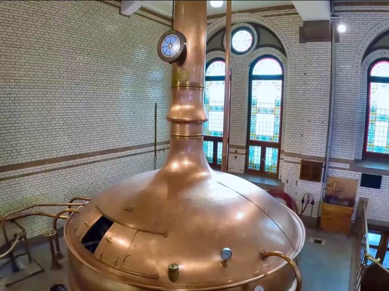 The interior of the Heineken experience shows a large copper brew kettle under tall windows with stained glass highlights