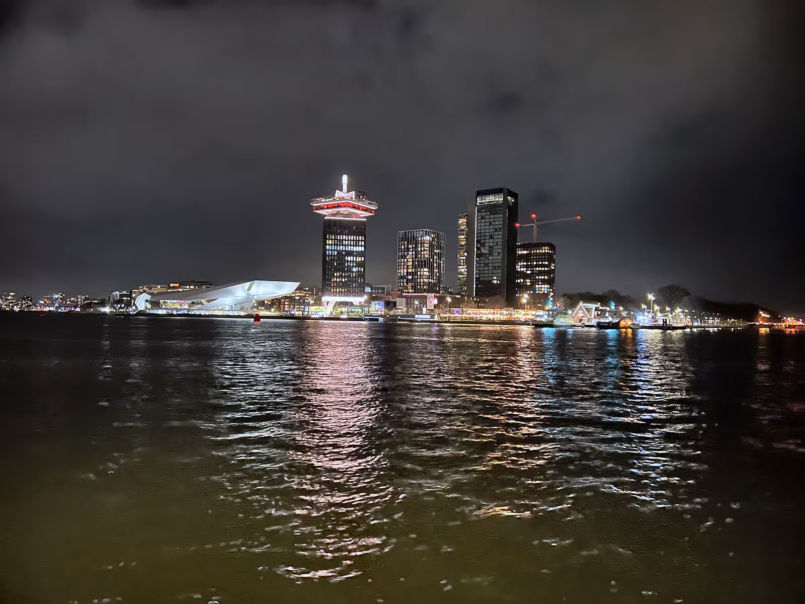 A nighttime view of Het IJ with city lights reflecting on the water and prominent buildings in the background