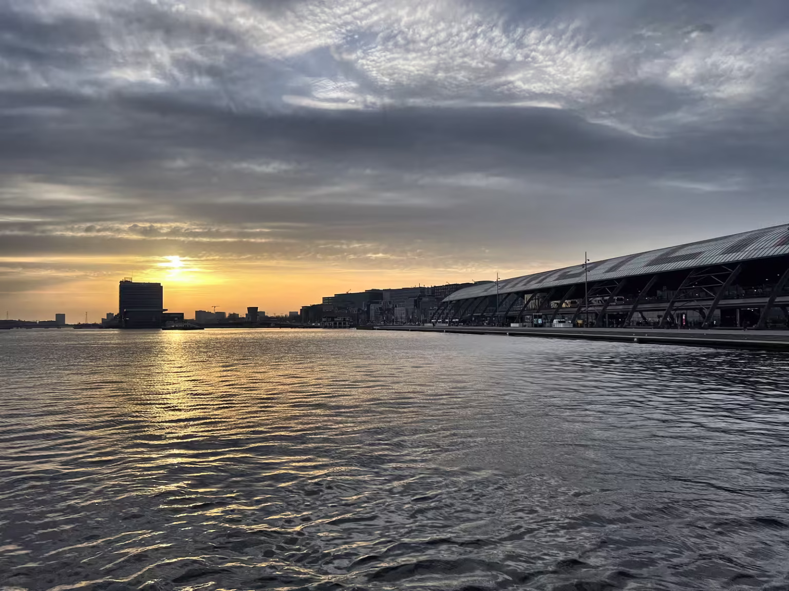 The tranquil waters of Het IJ reflect the sunset while buildings line the horizon creating a serene urban landscape