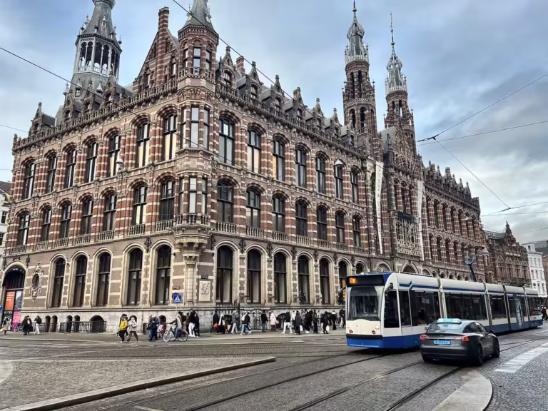 A historic building known as Magna Plaza showcases intricate architecture while a tram passes nearby and pedestrians walk