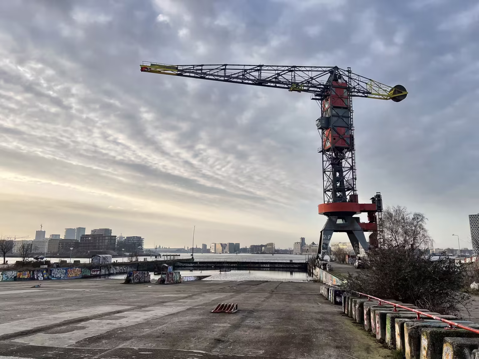 A large red and gray crane stands at the NDSM Wharf overlooking a calm waterway and the skyline beyond
