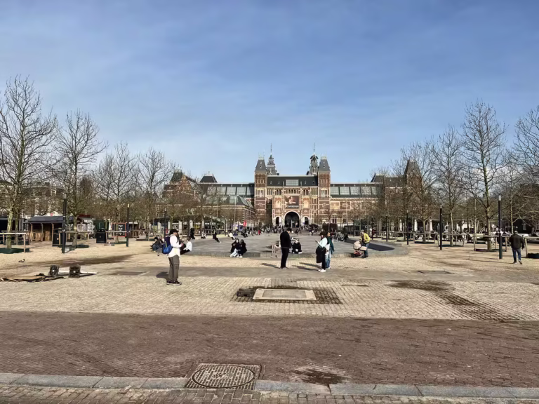A wide view shows the Rijksmuseum in amsterdam surrounded by trees and visitors enjoying the open space on a sunny day