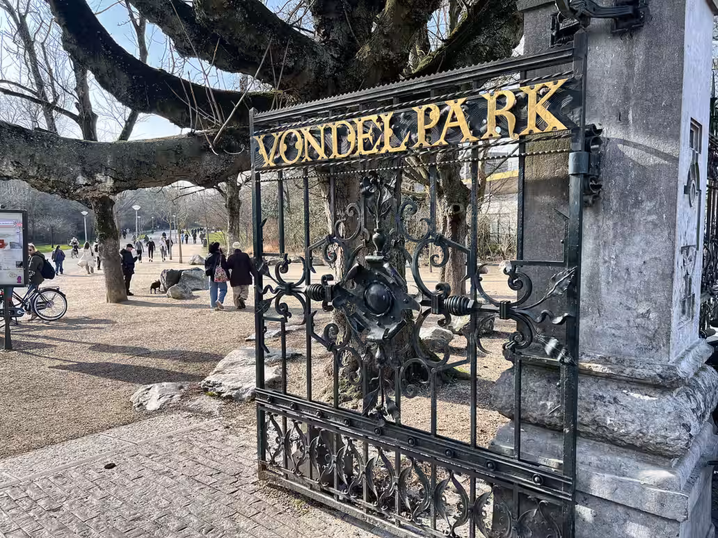 A decorative gate stands at the entrance to Vondelpark Amsterdam with people walking inside the park enjoying the scenery