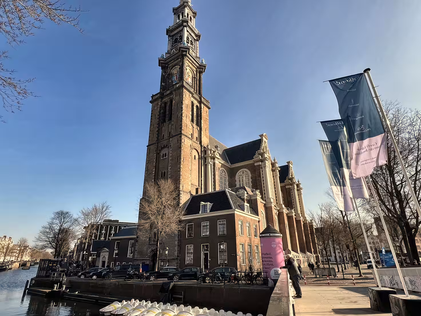 The Westerkerk stands majestically by a canal showcasing its tall tower and intricate architecture flanked by trees and city