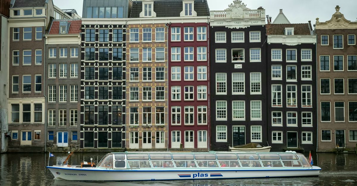 A passenger boat cruising on an Amsterdam canal in front of historic narrow canal houses, showcasing the city's traditional architecture and vibrant waterways.