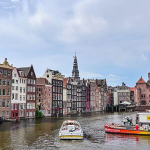 View of a canal in Amsterdam with typical Dutch canal houses lining the water and several small boats, including a canal tour boat, floating on the water under a cloudy sky.